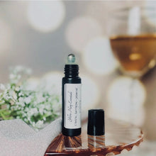 Load image into Gallery viewer, A black glass roll-on perfume oil bottle labeled 'Yellow Harp Essentials Crystal Saffron + Jasmine' stands on a wooden tray beside its cap, with a blurred glass of white wine and soft bokeh lights in the background