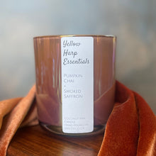 Load image into Gallery viewer, A candle in a iridescent, terra-cotta clay colored glass jar with a white label reading 'Yellow Harp Essentials' and 'Pumpkin Chai + Smoked Saffron,' styled on a wooden surface with draped rust-orange fabric against a soft gray-blue background.