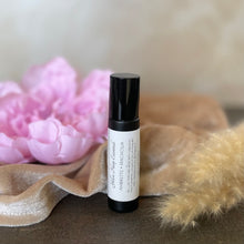 Load image into Gallery viewer, A small black glass cylindrical cosmetic rollerbottle with a white label reading 'Yellow Harp Essentials AMBRETTE + MAGNOLIA' perfume oil stands on a dark surface, styled with pink flowers, beige fabric, and soft tan decorative stems in the background.