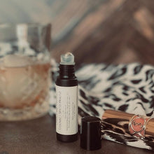 Load image into Gallery viewer, A small glass black roller bottle with a label reading 'Yellow Harp Essentials Cognac + Spiced Praline Rollerball Perfume Oil' stands open on a tabletop beside its cap, with a blurred drink and patterned fabric in the background.