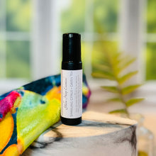 Load image into Gallery viewer, A small black glass roll-on perfume oil bottle with a white label reading 'Yellow Harp Essentials Sparkling Citrus + Green Tea' stands on a stone slab outdoors, with a colorful fabric and blurred greenery in the background.