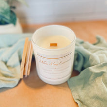 Load image into Gallery viewer, White candle in a textured container with a wooden wick on a light surface, surrounded by green fabric.