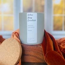 Load image into Gallery viewer, Gray glass candle jar with cork lid and white label that says “yellow harp essentials smoked lavender and amber coconut wax candle“ on wood table with full colored leaves out a back window
