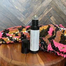 Load image into Gallery viewer, A small black glass roll-on bottle with a black cap placed beside it, set on a wooden surface in front of a sequined fabric. The bottle label reads 'Yellow Harp Essentials Honeyed Cinnamon + Osmanthus perfume oil'.
