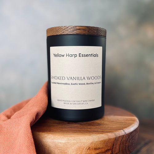 Candle labeled 'Yellow Harp Essentials Smoked Vanilla Woods' on a wooden surface with a blurred background
