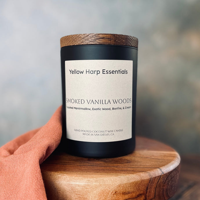 Candle labeled 'Yellow Harp Essentials Smoked Vanilla Woods' on a wooden surface with a blurred background