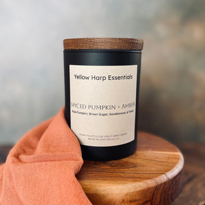 Candle labeled 'Yellow Harp Essentials Spiced Pumpkin + Amber' on a wooden surface with a blurred background