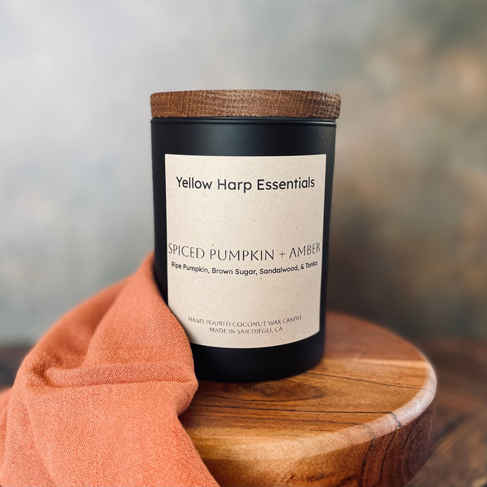 Candle labeled 'Yellow Harp Essentials Spiced Pumpkin + Amber' on a wooden surface with a blurred background