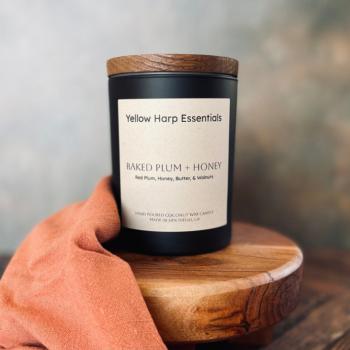 black candle jar with wooden lid and label from yellow harp essentials with baked plum and honey scent.