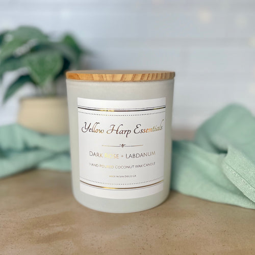 Candle with a wooden lid and 'Yellow Harp Essentials' label on a surface with greenery in the background