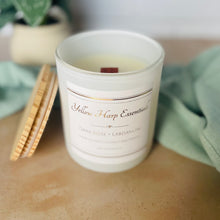 Load image into Gallery viewer, White candle in a glass jar with a label on a wooden surface, surrounded by greenery.
