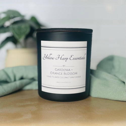 black glass jar with Yellow Harp Essentials on label and gardenia + orange blossom scent with greenery in background