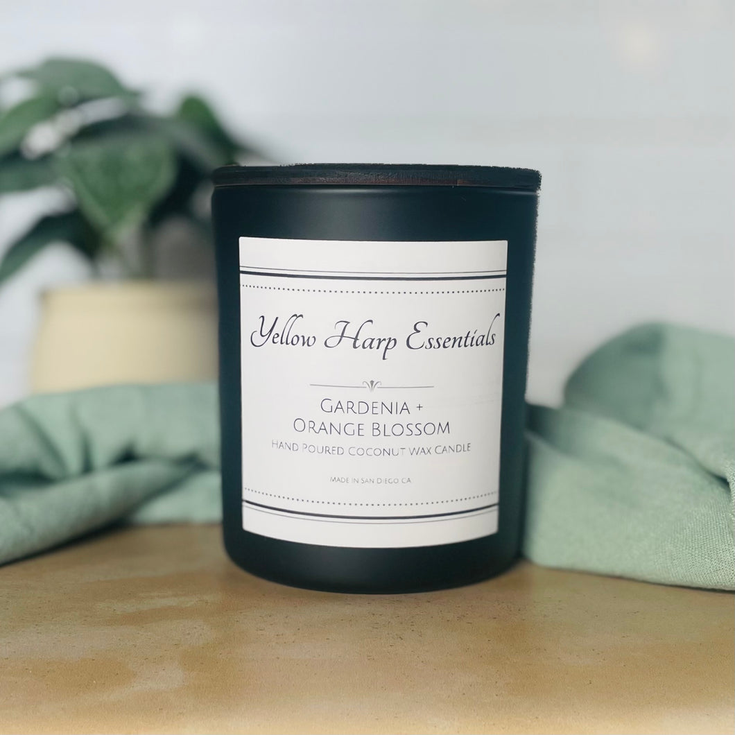 black glass jar with Yellow Harp Essentials on label and gardenia + orange blossom scent with greenery in background