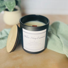 Load image into Gallery viewer, Candle in a black container with a label on a wooden surface, surrounded by greenery.
