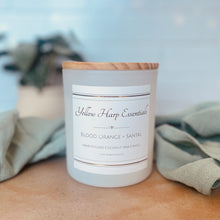 Load image into Gallery viewer, Candle with a wooden lid and 'Yellow Harp Essentials' label on a surface with a blurred background
