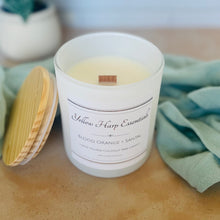 Load image into Gallery viewer, Candle with a wooden wick and white label on a wooden surface, surrounded by greenery.
